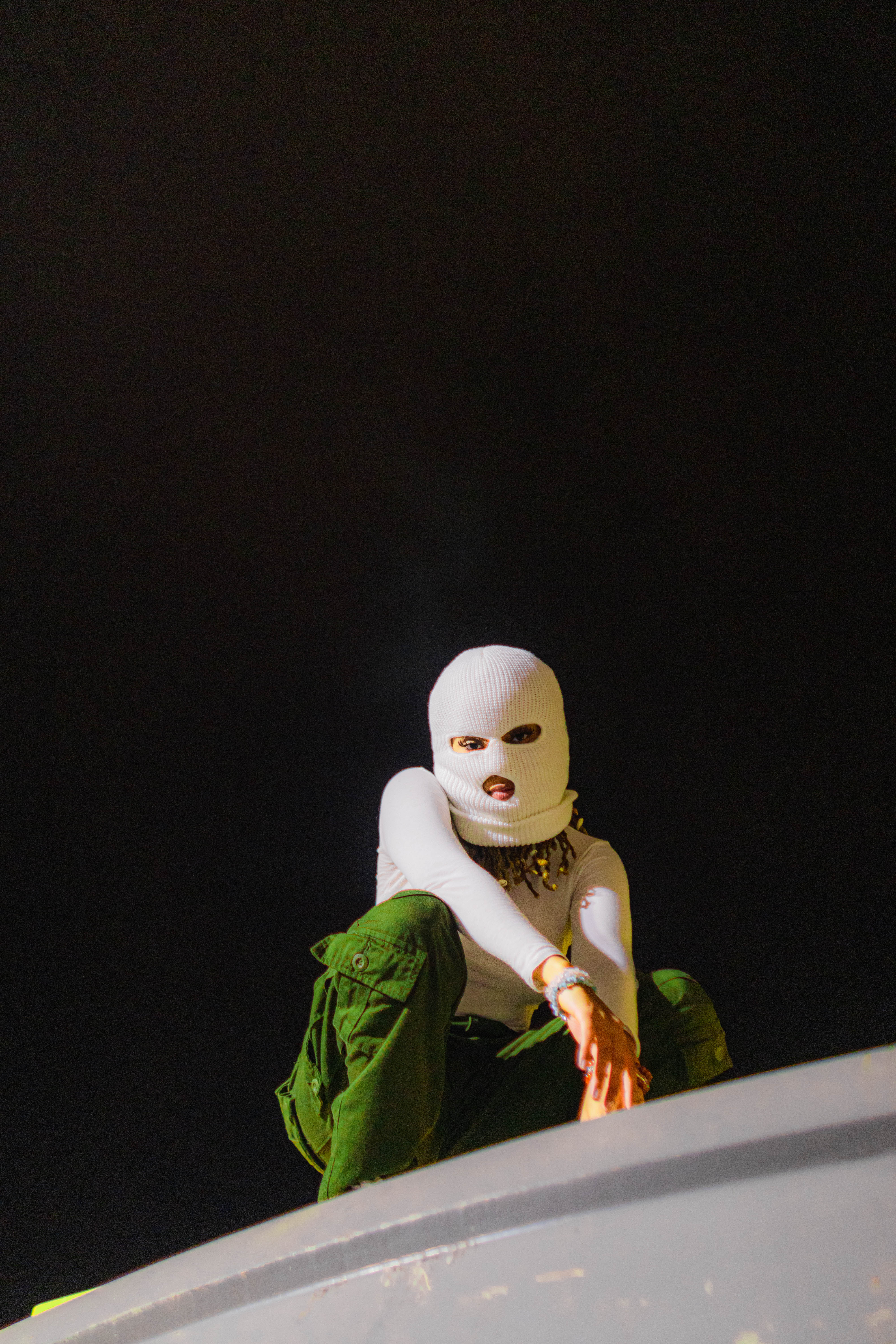 photograph shot from below of a person in a face-mask, 
    kneeling near the edge of a building and looking down at the camera